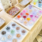 Bunte Perlen in verschiedenen Farben und Materialien, sortiert in kleinen Glasschälchen auf Tabletts. Im Hintergrund weitere Schalen mit Buchstabenperlen und farbigen Rocailles. Die Auswahl ist Teil des DIY Perlen Workshops bei Rosemary & Olive in Wuppertal 