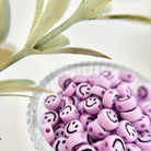 Nahaufnahme von fliederfarbenen Smileys-Perlen mit schwarzen Gesichtern in einer geriffelten Glasschale, perfekt für Freundschaftsarmbänder