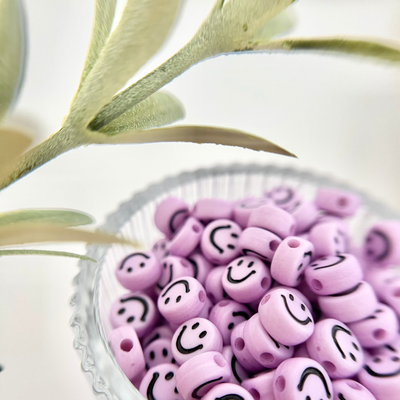 Nahaufnahme von fliederfarbenen Smileys-Perlen mit schwarzen Gesichtern in einer geriffelten Glasschale, perfekt für Freundschaftsarmbänder