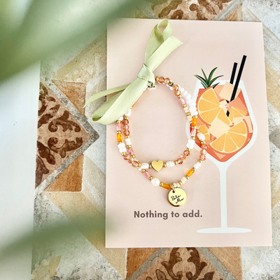 Zwei Perlenarmbänder auf illustrierten Cocktail-Postkarte mit Orangenmotiv und grüner Schleife. Geschenkfertiges Sommerduo mit warmen Farbtönen und Herz-Detail – ideal für Freundinnen. Typisches Aperol Armband. 