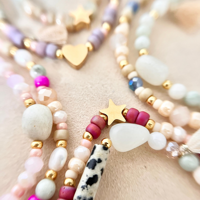 Nahaufnahme mehrerer selbst gestalteter Perlenarmbänder mit unterschiedlich großen Naturstein- und Glasperlen in Creme-, Rosé-, Beere- und Minttönen. Zu sehen sind goldfarbene Edelstahl-Zwischenelemente in Stern- und Herzform, ein schwarz-weiß gemusterter Naturstein sowie matt schimmernde Perlen, arrangiert auf hellem Stoff.