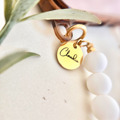 Detailaufnahme eines goldenen Gravur Anhängers mit Namen an einer Handykette mit weißen Perlen als personalisierbares Smartphone Accessoire für Alltag und Geschenk