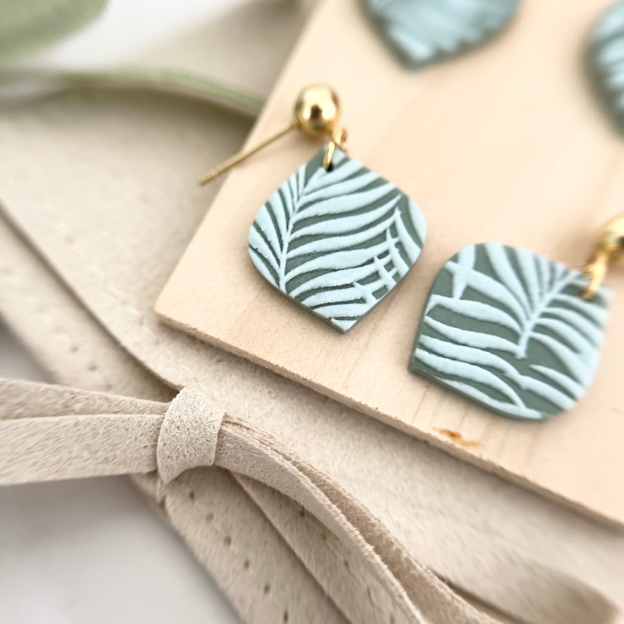 Nahaufnahme der Ohrringe HOJA aus Polymer Clay, goldfarben oder silberfarben, mit filigraner Palmenblatt-Prägung und Edelstahl-Stecker