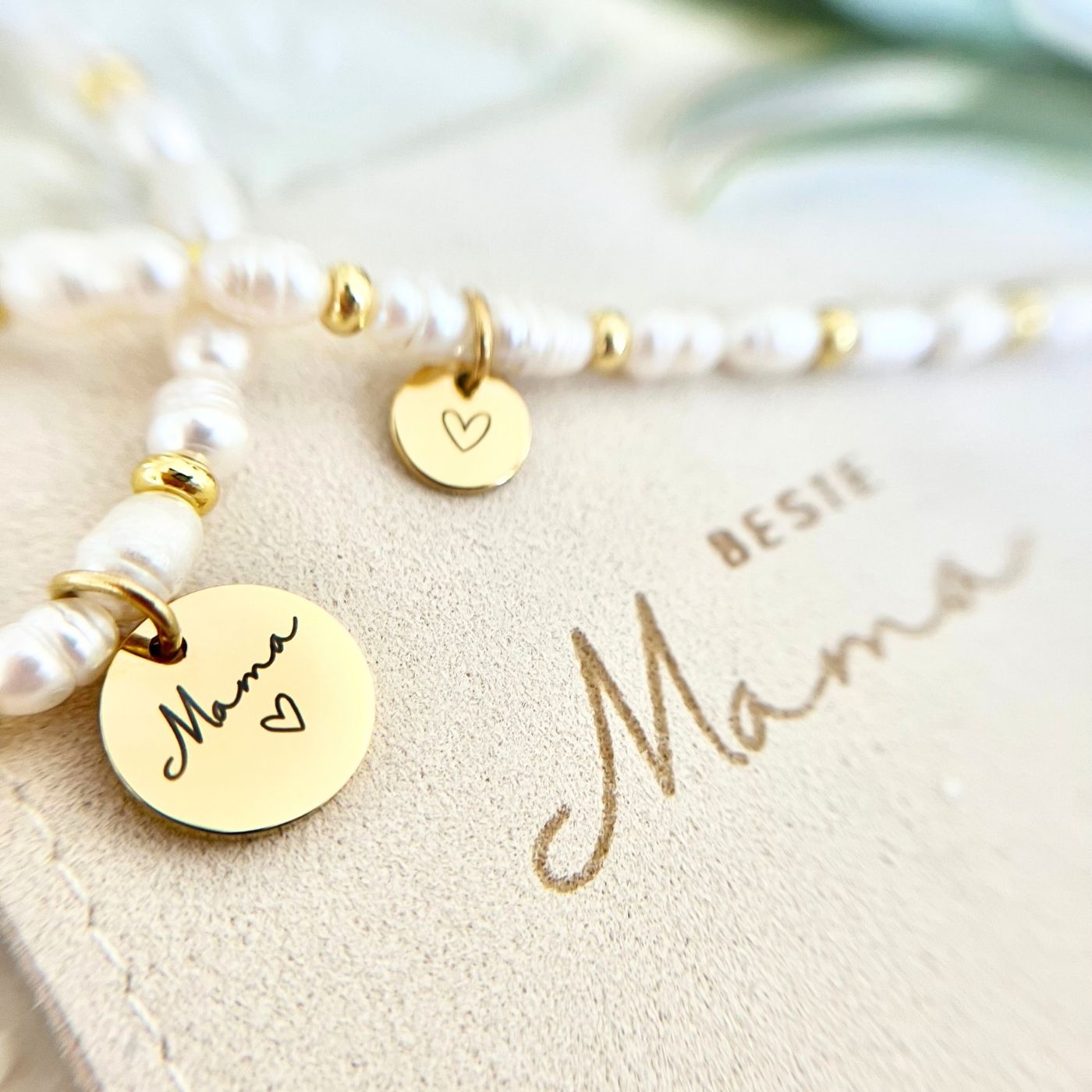 Detailaufnahme von zwei Gravur-Anhängern aus Edelstahl in Gold mit Aufschrift „Beste Mama“ und Herzsymbol am Süßwasserperlen-Armband.