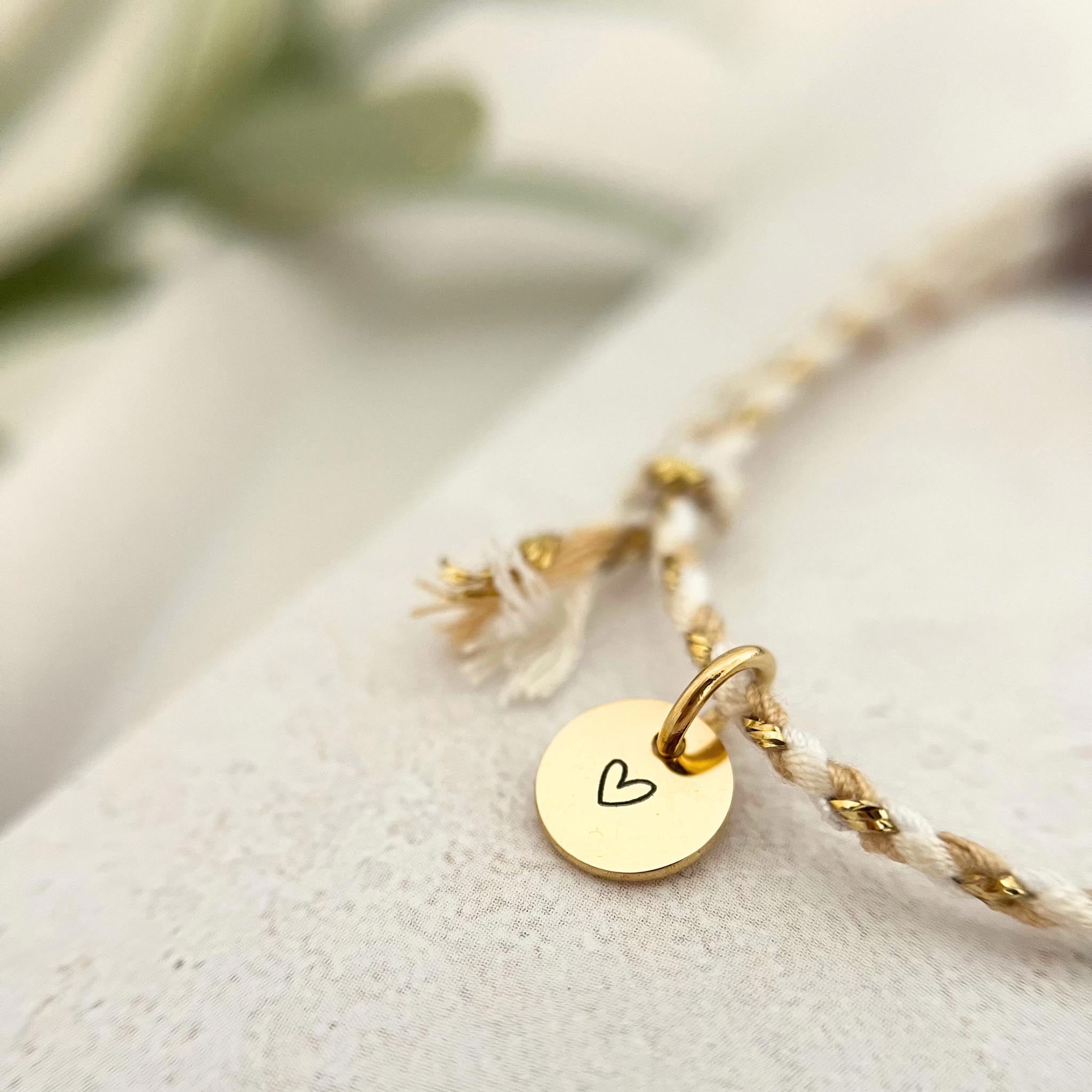 Nahaufnahme des goldenen Edelstahl-Anhängers mit Herzgravur auf dem Armband VINYOLA, kombiniert mit zarter Baumwollkordel in Weiß und Beige