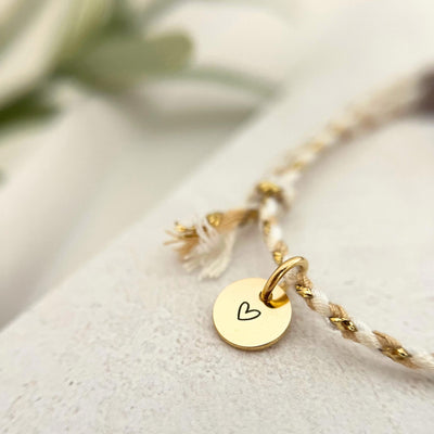 Nahaufnahme des goldenen Edelstahl-Anhängers mit Herzgravur auf dem Armband VINYOLA, kombiniert mit zarter Baumwollkordel in Weiß und Beige