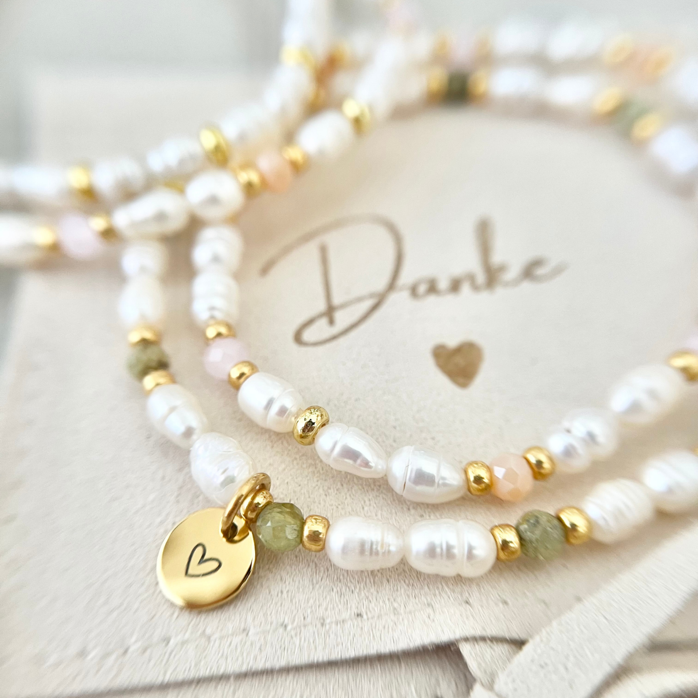 Handgemachtes Perlenarmband mit weißen Süßwasserperlen, grünen Natursteinen und goldenen Details. Der vergoldete Herzanhänger liegt auf einer Schmucktasche mit „Danke“-Aufschrift – stilvolles Dankeschön-Geschenk für Mama, Freundin oder Trauzeugin.