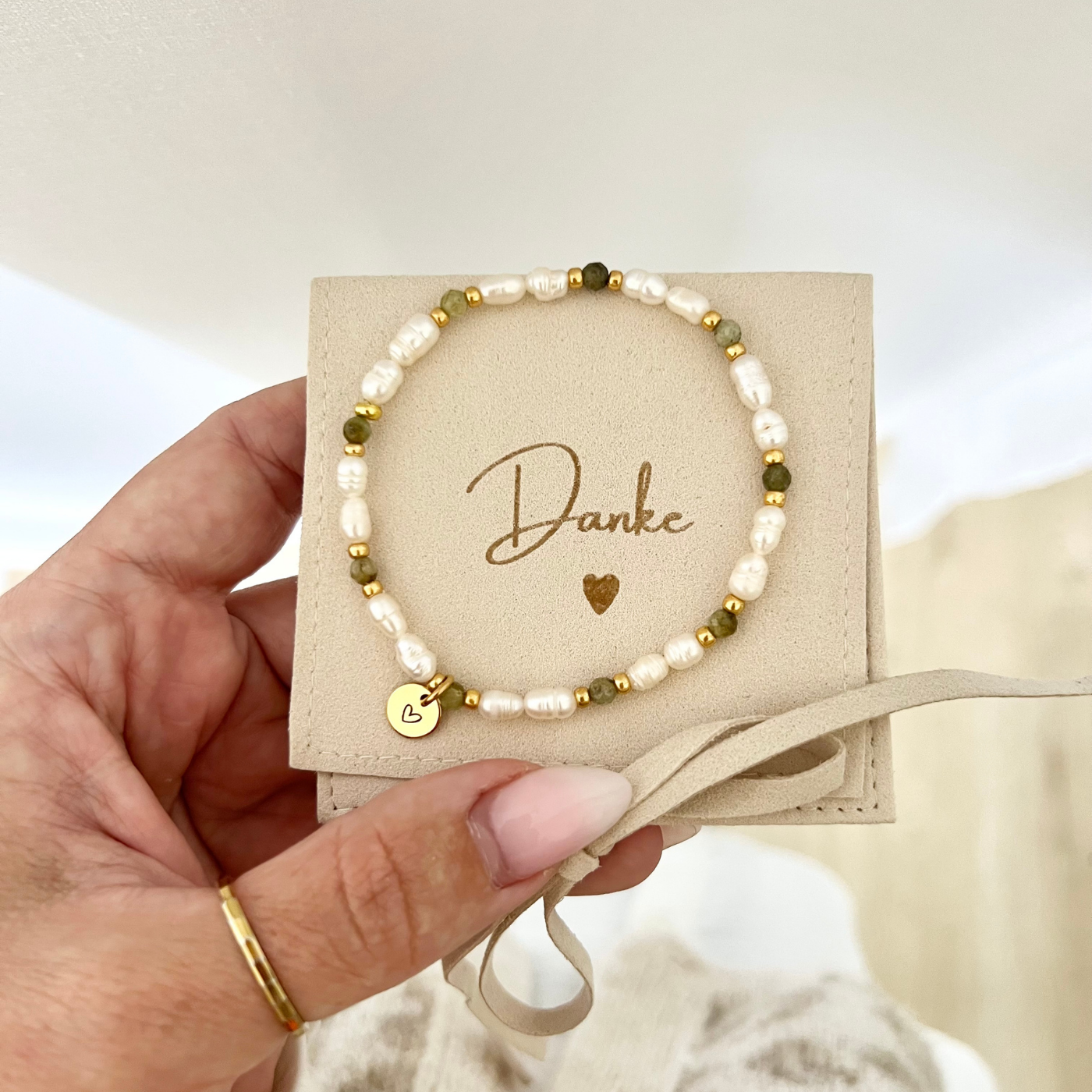 Handgemachtes Armband mit weißen Süßwasserperlen, grünen Natursteinperlen und goldenen Details, präsentiert auf einer Schmucktasche mit „Danke“-Aufschrift. Ein vergoldeter Herzanhänger macht es zum idealen Dankeschön-Geschenk für die Trauzeugin nach der Hochzeit.
