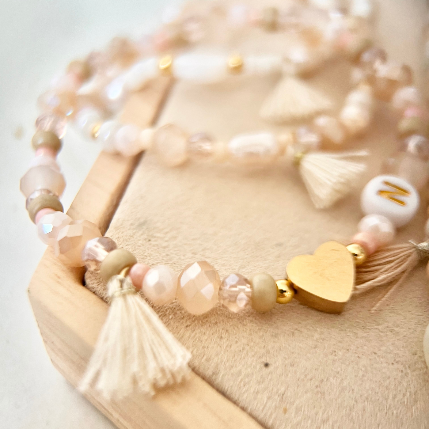 Nahaufnahme eines selbst gestalteten Perlenarmbands mit Glasperlen in Rosé- und Cremetönen, goldfarbenem Herz-Anhänger, Quaste und Buchstabenperle auf heller Holzoberfläche.