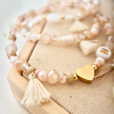 Nahaufnahme eines selbst gestalteten Perlenarmbands mit Glasperlen in Rosé- und Cremetönen, goldfarbenem Herz-Anhänger, Quaste und Buchstabenperle auf heller Holzoberfläche.