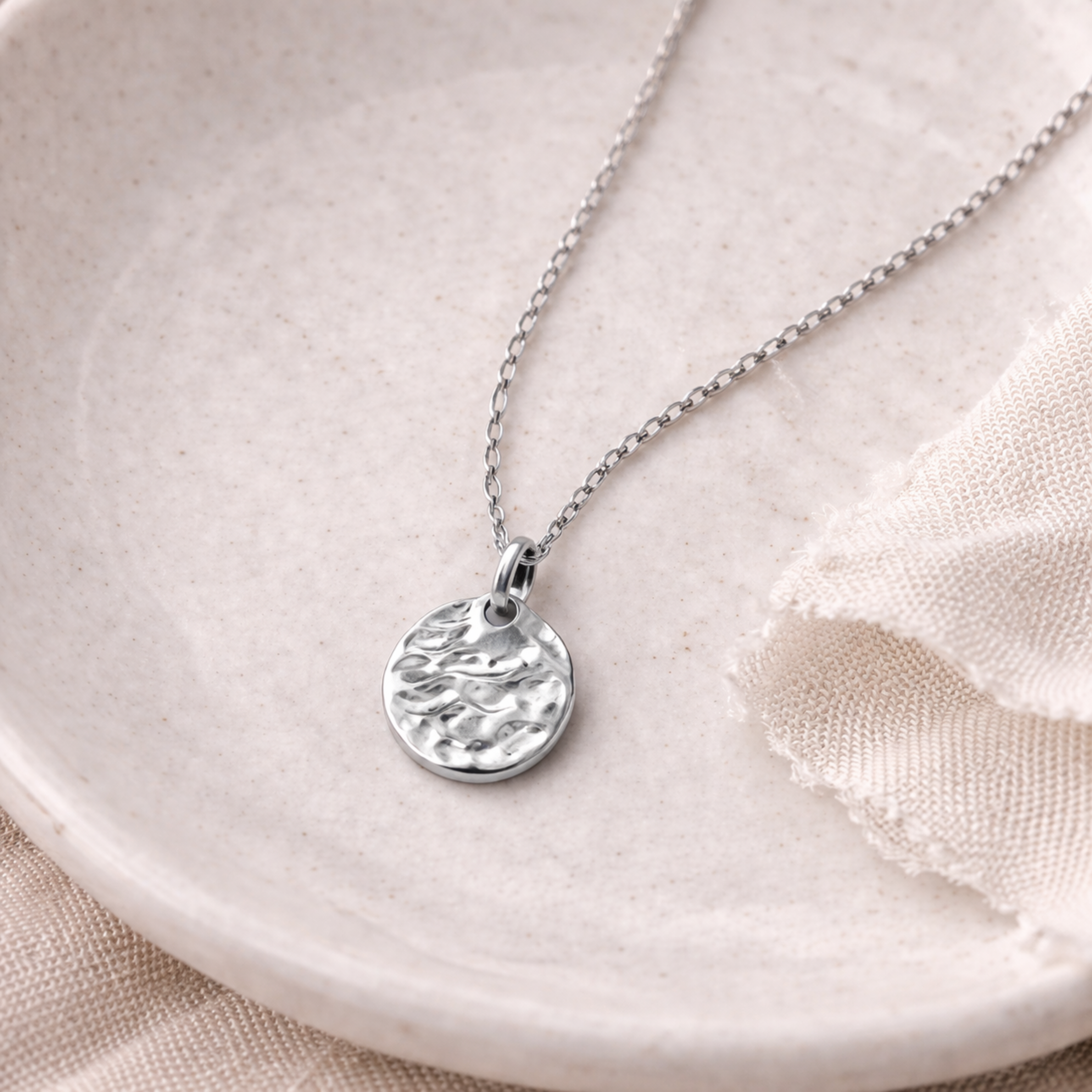 Silberne Edelstahl Halskette mit kleinem gehämmerten Anhänger auf heller Keramikschale und beigem Leinenstoff, Detailaufnahme Schmuck für Frauen