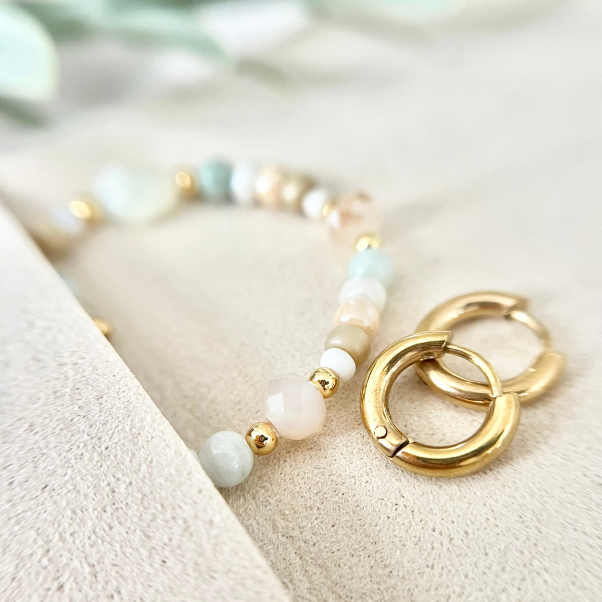 Nahaufnahme eines Armbands mit kleinen runden Perlen in Weiß, Beige und Mint sowie goldfarbenen Edelstahl Creolen, liegend auf einem hellen Schmuckbeutel.