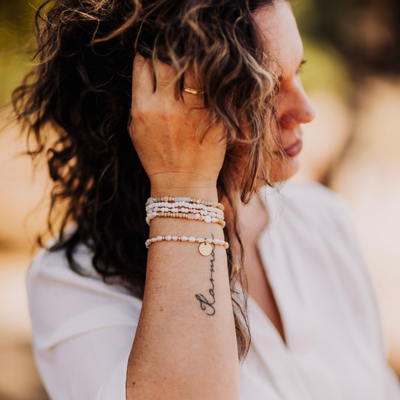 Frau mit lockigen Haaren und weißer Bluse trägt mehrere filigrane Armbänder aus Perlen und vergoldeten Details am Handgelenk. Ein runder Anhänger mit Gravur fällt ins Auge, daneben sichtbar ein Tattoo mit dem Schriftzug „Karma“ – stilvolles Sommer-Accessoire im Boho-Look.