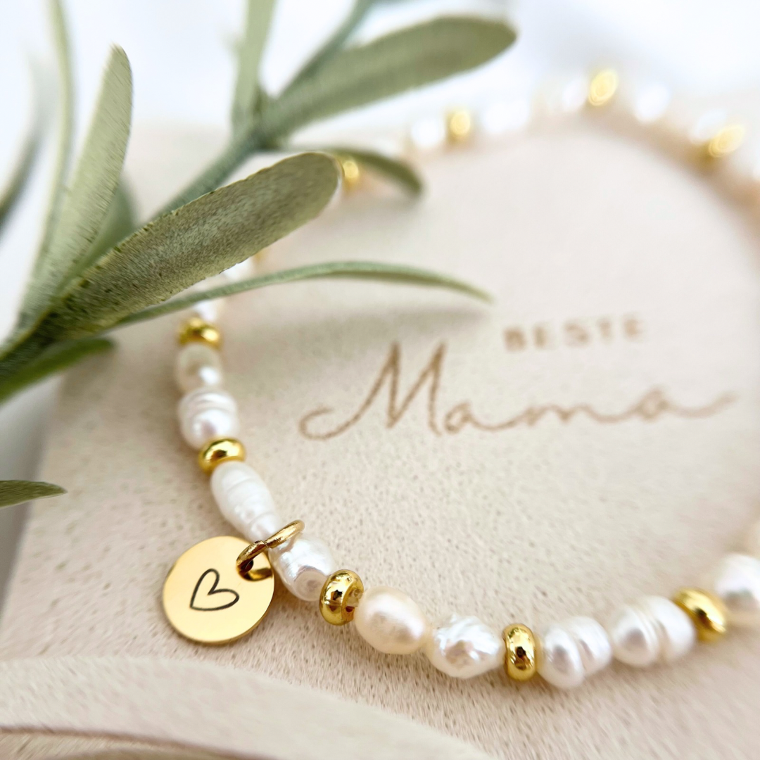 Nahaufnahme eines zarten Armbands aus unregelmäßigen Süßwasserperlen und goldenen Zwischenperlen, mit rundem goldfarbenem Anhänger mit eingraviertem Herz. Im Hintergrund steht auf der Schmuckverpackung die Gravur „Beste Mama“.