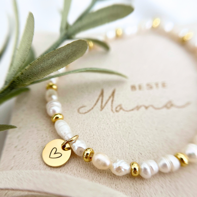 Nahaufnahme eines zarten Armbands aus unregelmäßigen Süßwasserperlen und goldenen Zwischenperlen, mit rundem goldfarbenem Anhänger mit eingraviertem Herz. Im Hintergrund steht auf der Schmuckverpackung die Gravur „Beste Mama“.