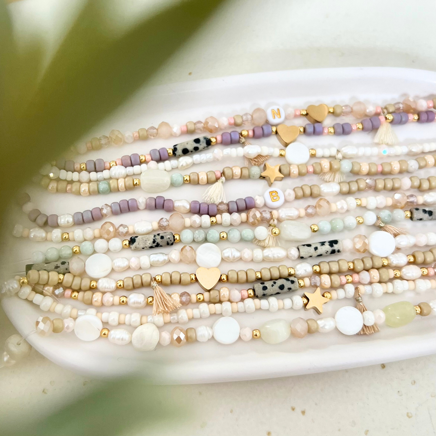 Mehrere handgemachte Beispiele für Perlenarmbänder nebeneinander auf einem weißen Teller, mit Naturstein-, Glas- und Süßwasserperlen in Creme-, Rosé- und Fliedertönen, goldfarbenen Herz- und Sternanhängern sowie kleinen Quasten.