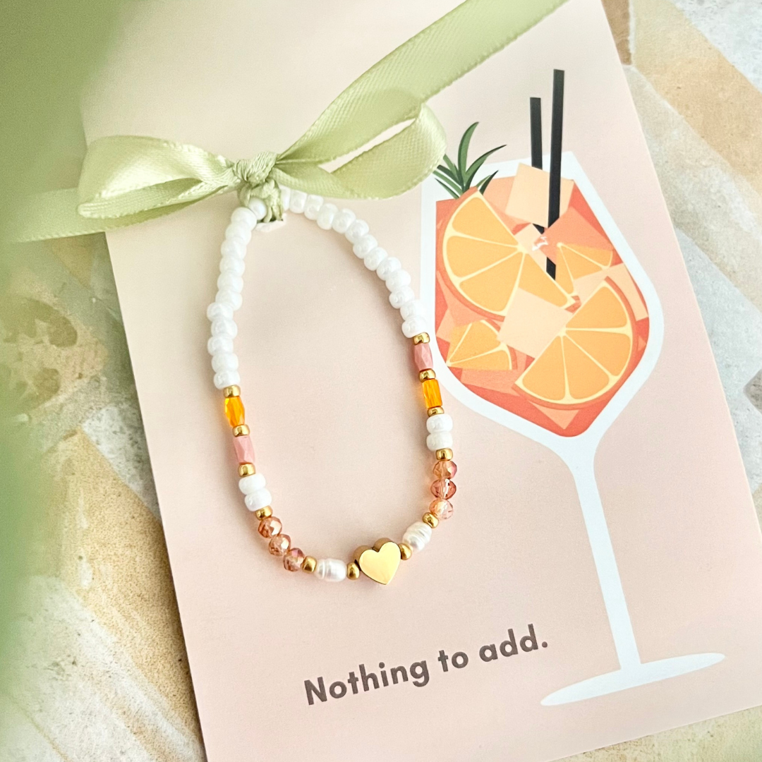 Perlenarmband in warmen Orangetönen und Weiß mit goldener Herz Perle auf einer rosa Postkarte mit Getränkemotiv und heller Satin-Schleife befestigt.