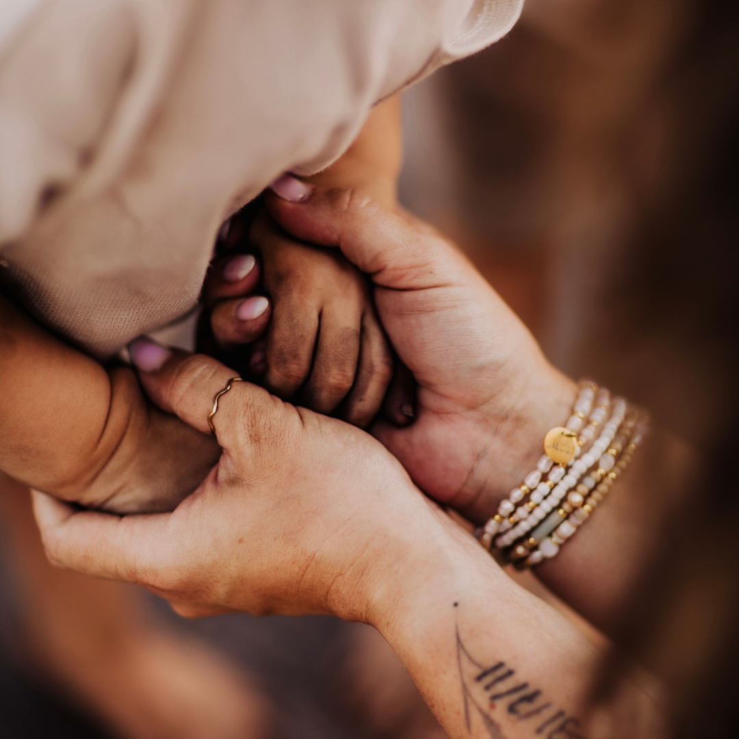 Inniger Moment: Eine Mutter hält liebevoll die Hand ihres Kindes. Am Handgelenk trägt sie mehrere zarte Perlenarmbänder mit goldenen Details und einem gravierten Anhänger – Symbol für Verbundenheit und Liebe.