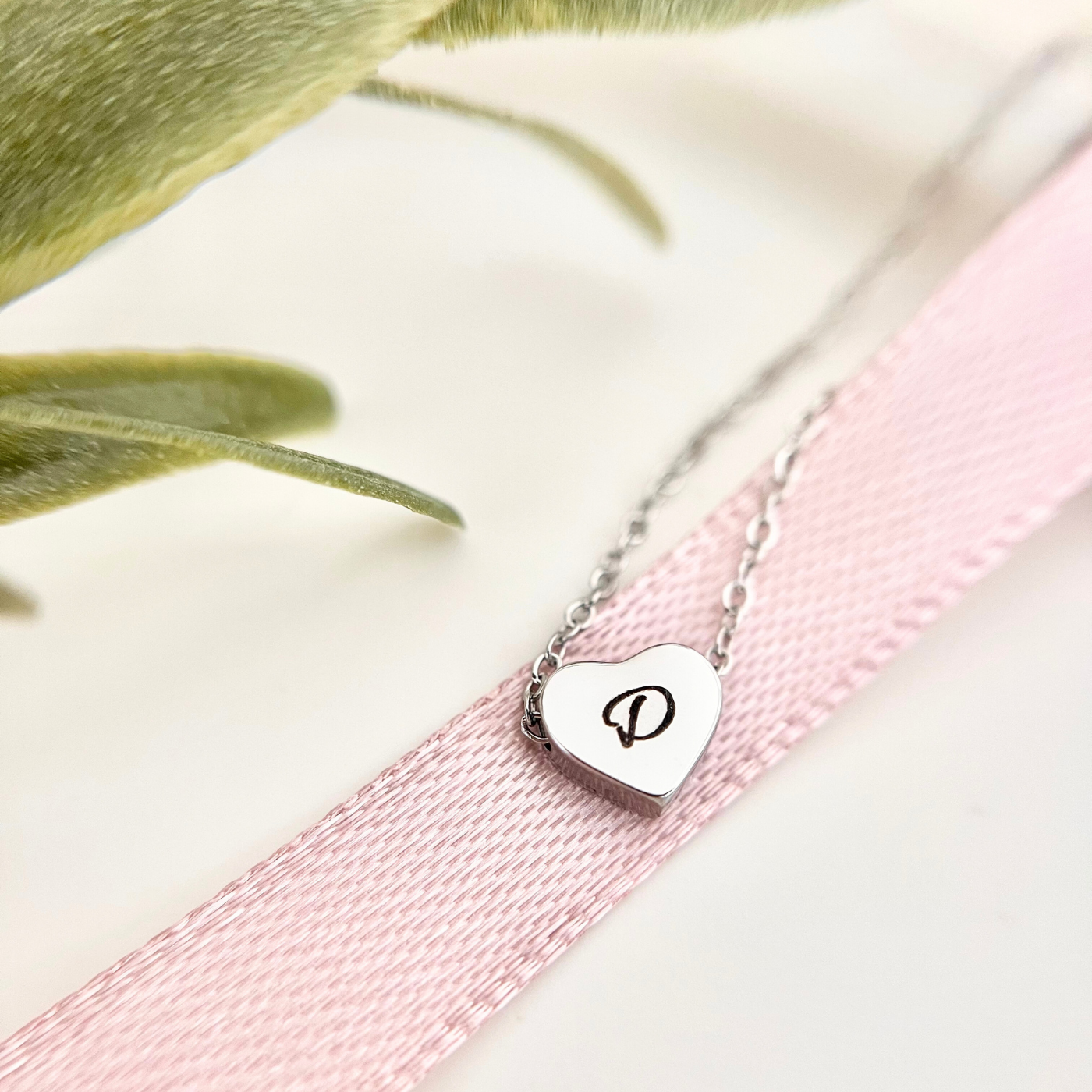Nahaufnahme einer filigranen Edelstahlkette mit kleinem Herzanhänger, personalisiert mit dem Buchstaben „D“. Die Kette liegt auf einem rosafarbenen Stoffband, daneben sind grüne Zweige zu sehen. Das Schmuckstück dient als persönliches Geschenk oder liebevolle Erinnerung für besondere Momente.