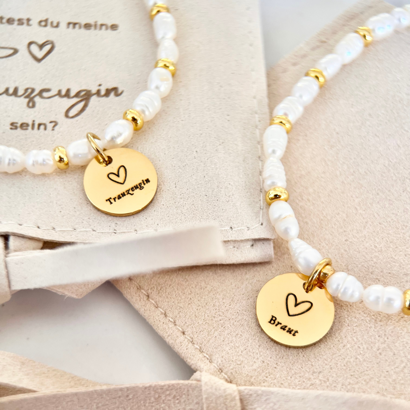 Zwei feine Perlenarmbänder mit goldfarbenen Gravur Anhängern „Braut“ und „Trauzeugin“, arrangiert auf beigen Schmuckbeuteln.
