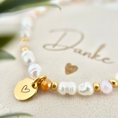 Detailaufnahme eines handgemachten Armbands mit weißen Süßwasserperlen, apricot farbenen Glasperlen, goldfarbenen Zwischenelementen und rundem Edelstahl-Anhänger mit eingraviertem Herz, dekorativ auf Schmuckbeutel mit der Aufschrift „Danke“.