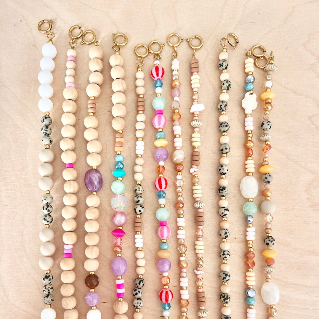 Draufsicht auf mehrere im Handyketten Workshop gefertigte kurze Perlen-Handyketten in unterschiedlichen Designs mit Holzperlen, Acrylperlen, Glasperlen und Natursteinperlen in Beige-, Rosa-, Türkis-, Orange- und Schwarz-Weiß-Tönen sowie goldfarbenen Verschlüssen auf hellem Holzuntergrund.