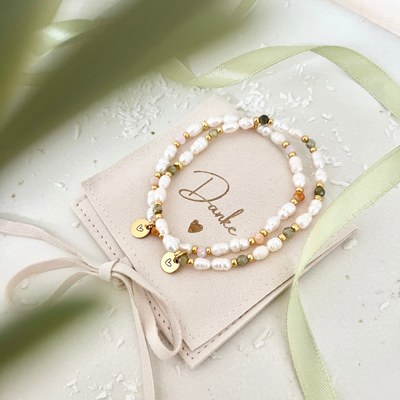 Zwei elegante Armbänder aus weißen Süßwasserperlen mit goldfarbenen Zwischenelementen und grünen sowie apricotfarbenen Glasperlen, verziert mit Herz-Anhängern aus Edelstahl. Präsentiert auf einem Schmuckbeutel mit der Aufschrift „Danke“, umgeben von grünem Satinband – stilvolles Weihnachtsgeschenk für Mama, Freundin oder Trauzeugin.