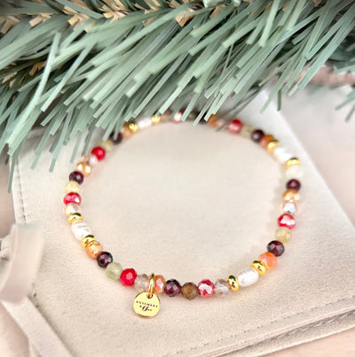 Weihnachtliches Perlenarmband NOELLE aus Naturstein, facettierten Glasperlen und weißen Süßwasserperlen in Rot, Weinrot, Grün, Apricot und Gold. Mit kleinem goldenen Logoanhänger, dekoriert auf einem cremefarbenen Schmuckbeutel unter Tannenzweigen.