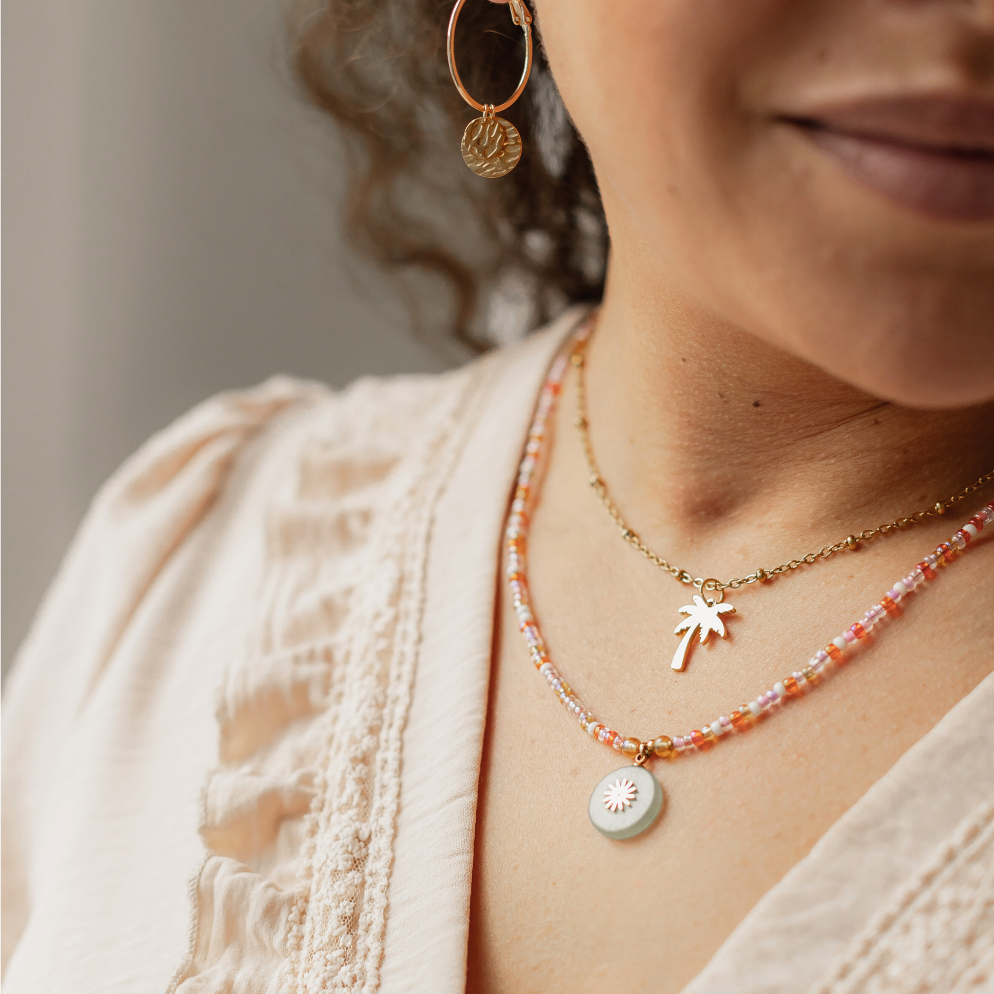 Nahaufnahme von Model trägt die Kette mit Naturstein-Anhänger mit Sonne darauf. Model trägt noch eine andere Kette mit Palmen Anhänger und einen goldenen Ohrring
Model trägt die Kette ARTÀ mit einem Naturstein-Sonnenanhänger, kombiniert mit einer Palmenanhänger-Kette und einem goldenen Ohrring, die den sommerlichen Look ergänzen.