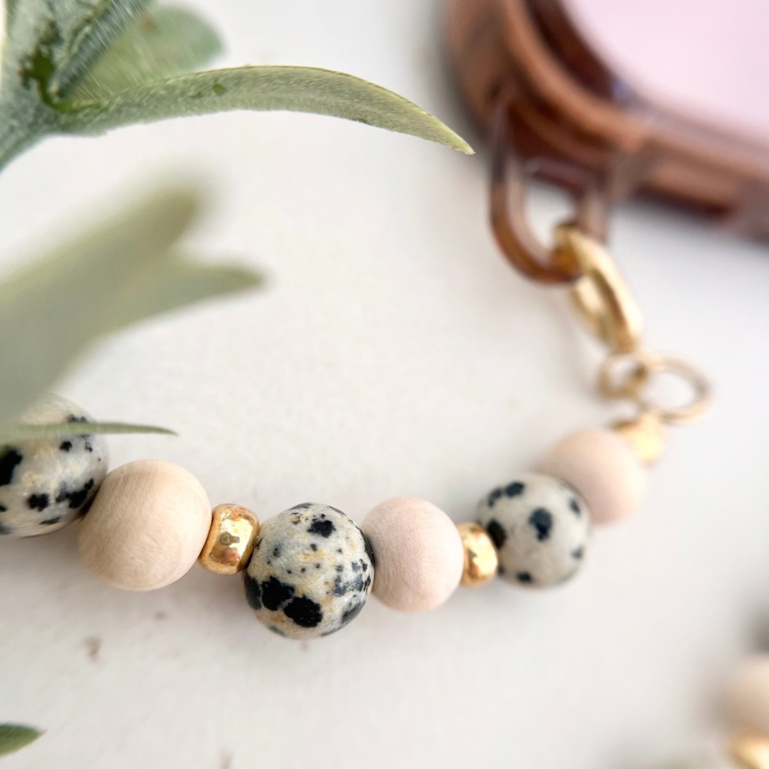 Nahaufnahme einer im Workshop gefertigten handgemachten Perlen-Handykette mit runden Holzperlen in Naturbeige, Dalmatiner Jaspis Natursteinperlen mit schwarzen Sprenkeln sowie kleinen goldfarbenen Metallelementen und Karabinerverschluss vor hellem Hintergrund mit Pflanze.