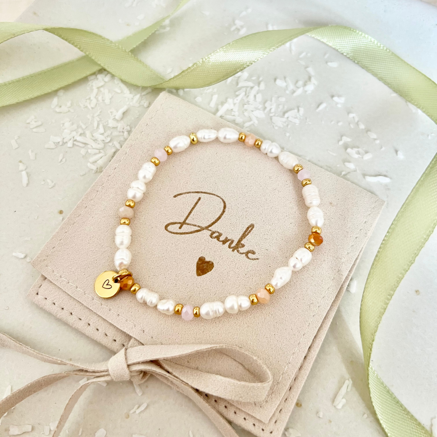 Elegantes Perlenarmband aus weißen Süßwasserperlen, kombiniert mit goldfarbenen Zwischenelementen und apricotfarbenen Glasperlen. Mit rundem Edelstahl-Anhänger mit Herzgravur, dekorativ auf beigem Schmuckbeutel mit der Aufschrift „Danke“, umgeben von grünem Satinband.