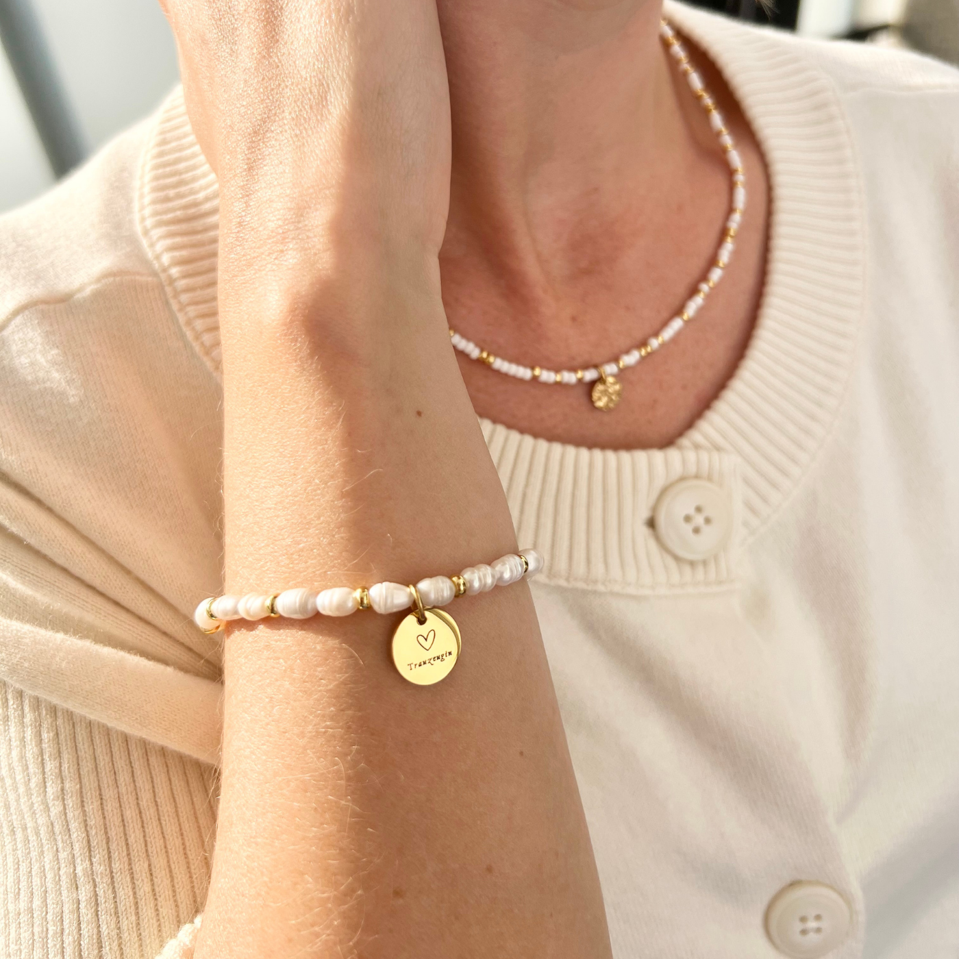 Nahaufnahme eines feinen Perlenarmbands aus weißen Süßwasserperlen mit rundem, goldenem Anhänger mit Gravur „Trauzeugin“, getragen am Handgelenk.
