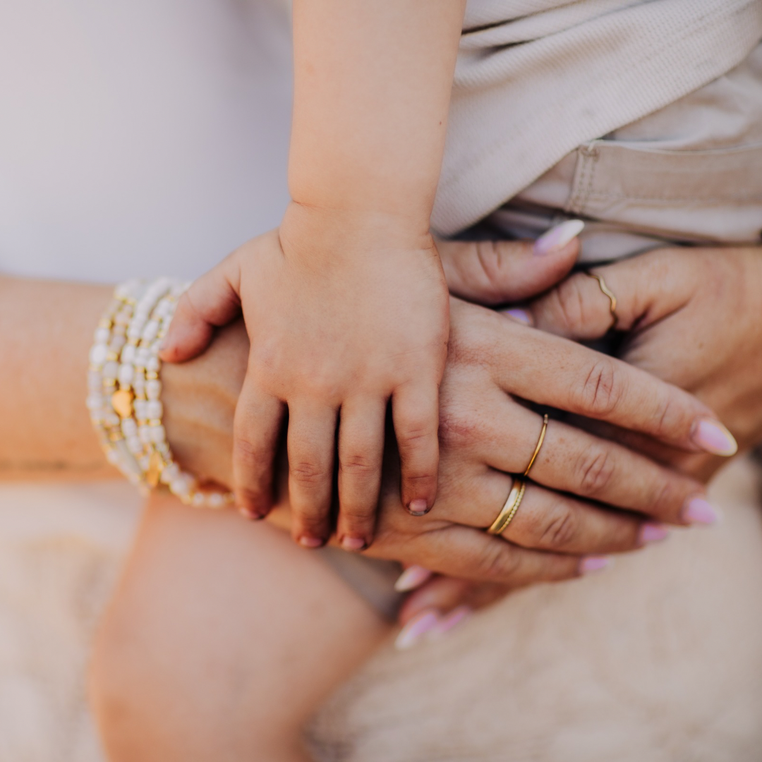 Nahaufnahme einer Kinderhand, die liebevoll auf den Händen einer Frau mit goldenen Ringen und weißen Perlenarmbändern liegt – symbolträchtiges Schmuckgeschenk für die Patentante