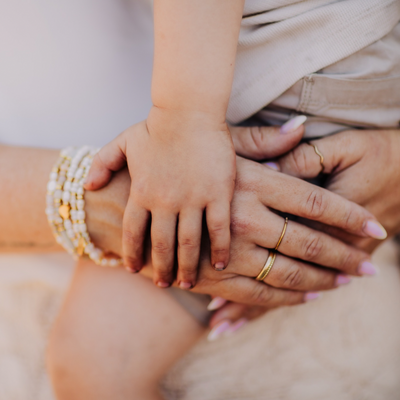 Nahaufnahme einer Kinderhand, die liebevoll auf den Händen einer Frau mit goldenen Ringen und weißen Perlenarmbändern liegt – symbolträchtiges Schmuckgeschenk für die Patentante