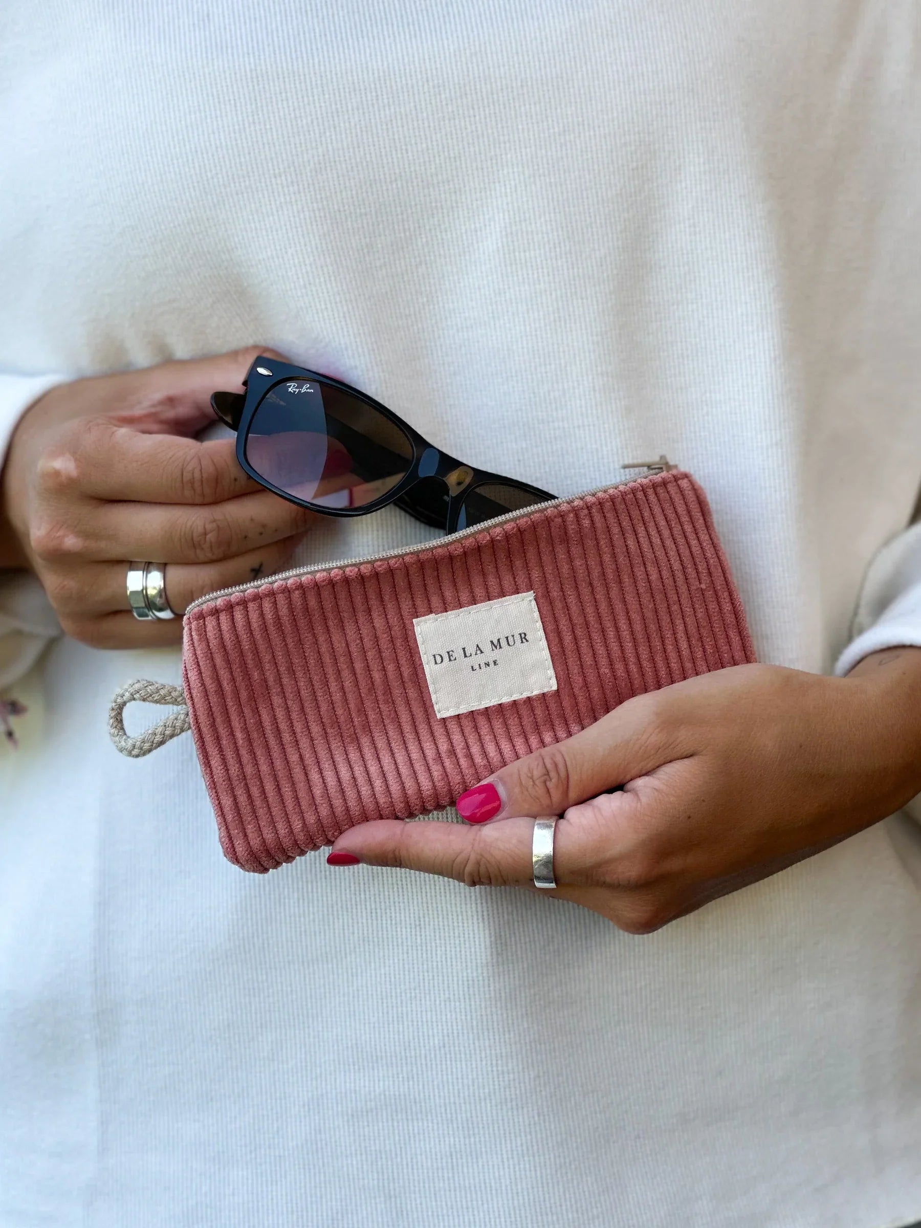 Eine Person in weißem Oberteil hält eine schwarze Sonnenbrille und ein korallfarbenes Cord-Brillenetui mit DE LA MUR Label. Die Hände sind leicht angewinkelt, die Fingernägel rosa lackiert.