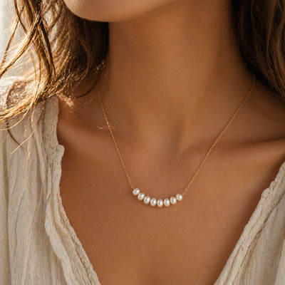 Goldene, filigrane Halskette mit kleinen Süßwasserperlen, getragen auf sonnengeküsster Haut im Boho-Stil; warmes Licht, natürlicher Look, Fokus auf Dekolleté und Schmuckstück.
