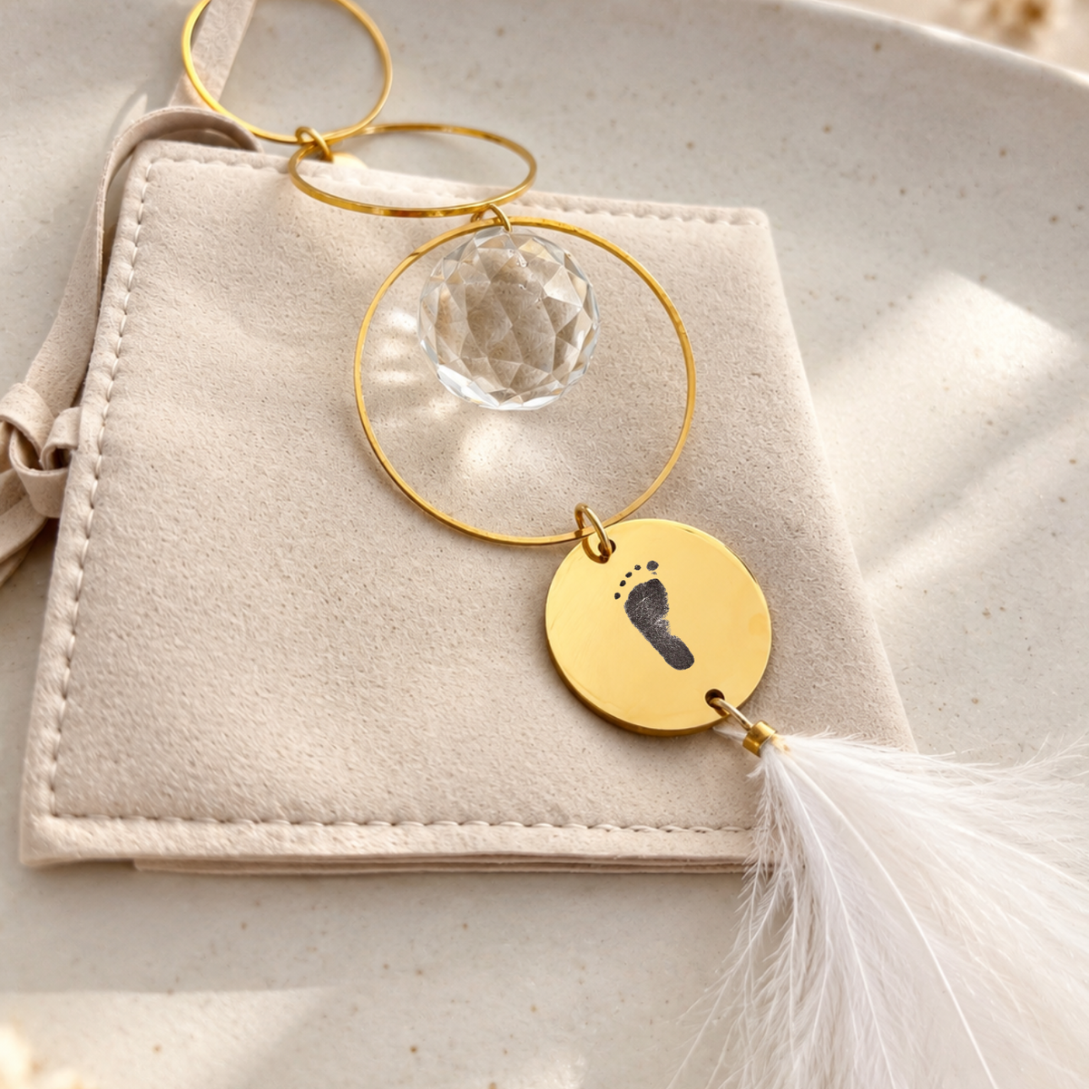 Goldener Sonnenfänger mit Kristallkugel, graviertem Baby-Fußabdruck und weißer Feder auf einem beigen Schmuckbeutel als Geschenk zur Geburt.