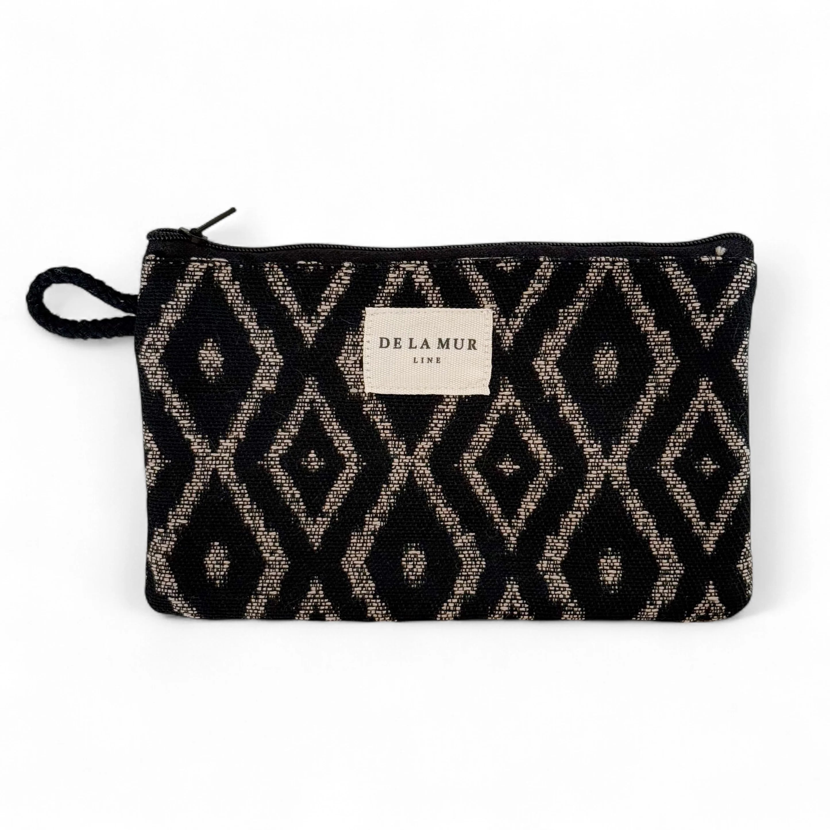 Rechteckige kleine Jacquard-Tasche von DE LA MUR mit grafischem Rautenmuster in Beige auf schwarzem Hintergrund, Reißverschluss und seitlicher Kordelschlaufe. Dient zur Aufbewahrung von Make-up, Pflegeprodukten oder Schmuck.
