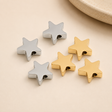 Goldene und silberne Sternperlen aus Edelstahl auf hellem beigen Hintergrund als Schmuckzubehör für Armbänder, Ketten und DIY-Schmuck.
