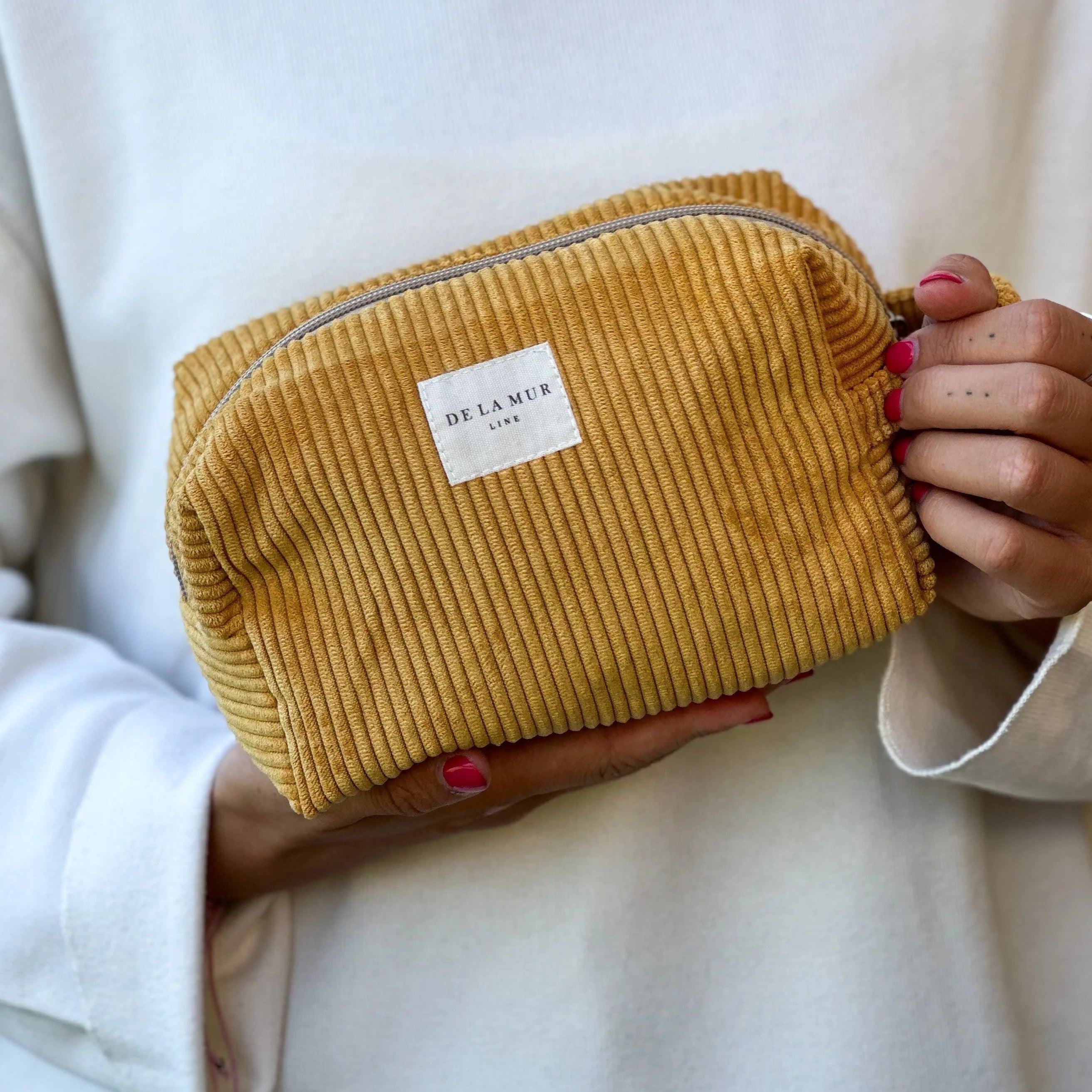 andgefertigte Beauty Bag Medium von DE LA MUR aus senfgelbem Cordstoff mit Rippenstruktur, rechteckige Form mit Reißverschluss und seitlicher Schlaufe, gehalten in Händen. Ideal zur Aufbewahrung von Kosmetik, Pflegeartikeln oder Schmuck auf Reisen.