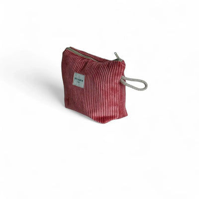 Eine kleine, korallenfarbene Cord-Tasche steht im Dreiviertelwinkel vor weißem Hintergrund. Seitlich befindet sich eine graue Schlaufe, oben ein Reißverschluss, vorne das DE LA MUR Label.