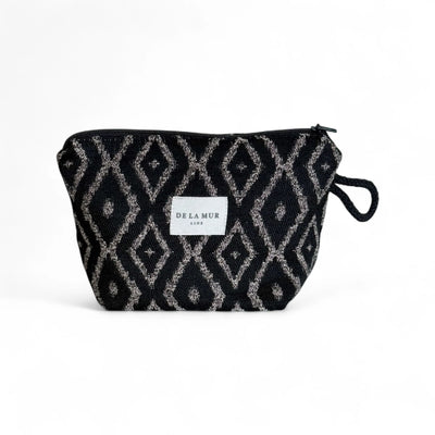 Eine rechteckige Kosmetiktasche mit schwarzem Jacquardstoff und beigefarbenem Rautenmuster steht im vor weißem Hintergrund. Oben ist ein schwarzer Reißverschluss sichtbar, an der Seite hängt eine kleine Schlaufe, vorne klebt das weiße DE LA MUR Label.