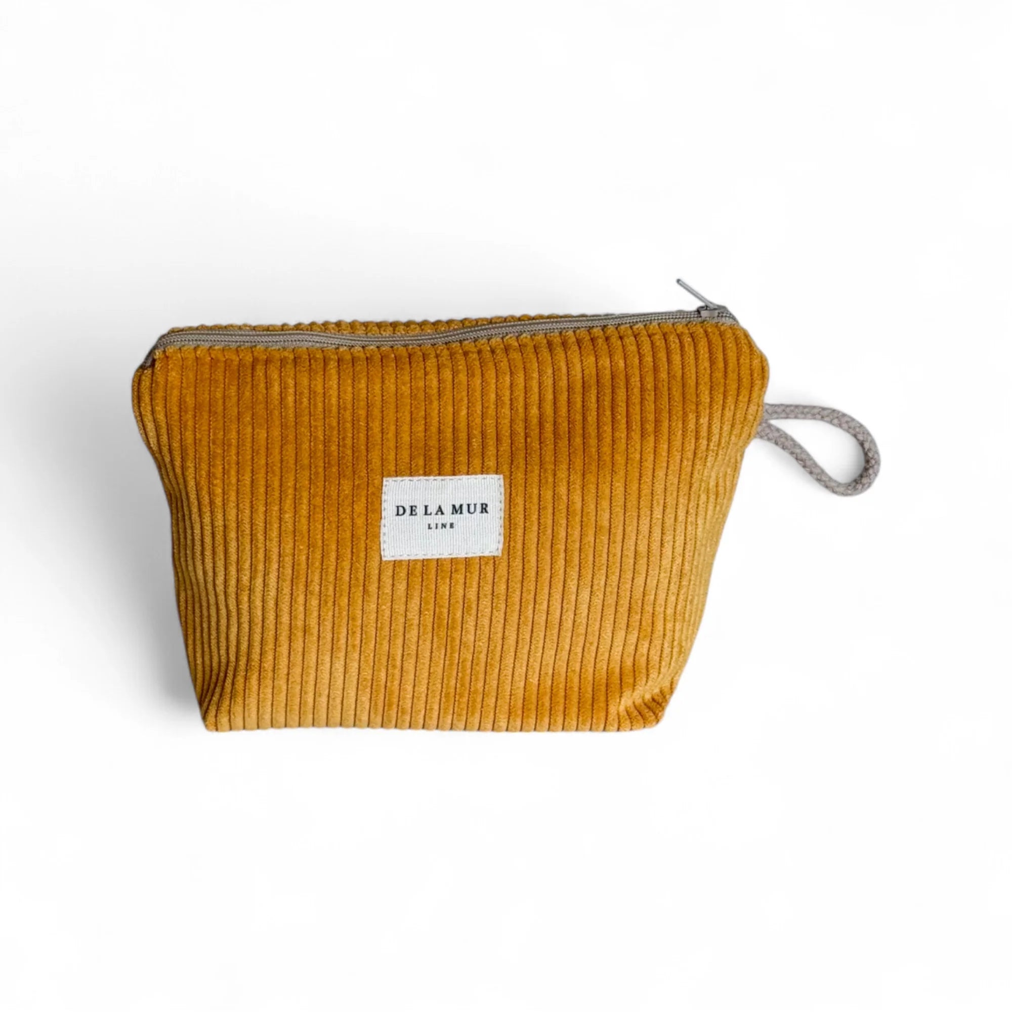 Eine senfgelbe Cord-Kosmetiktasche mit ausgeprägter Rippenstruktur liegt vor weißem Hintergrund. Ein grauer Reißverschluss verläuft oben, und mittig vorne ist ein weißes DE LA MUR Label aufgenäht.