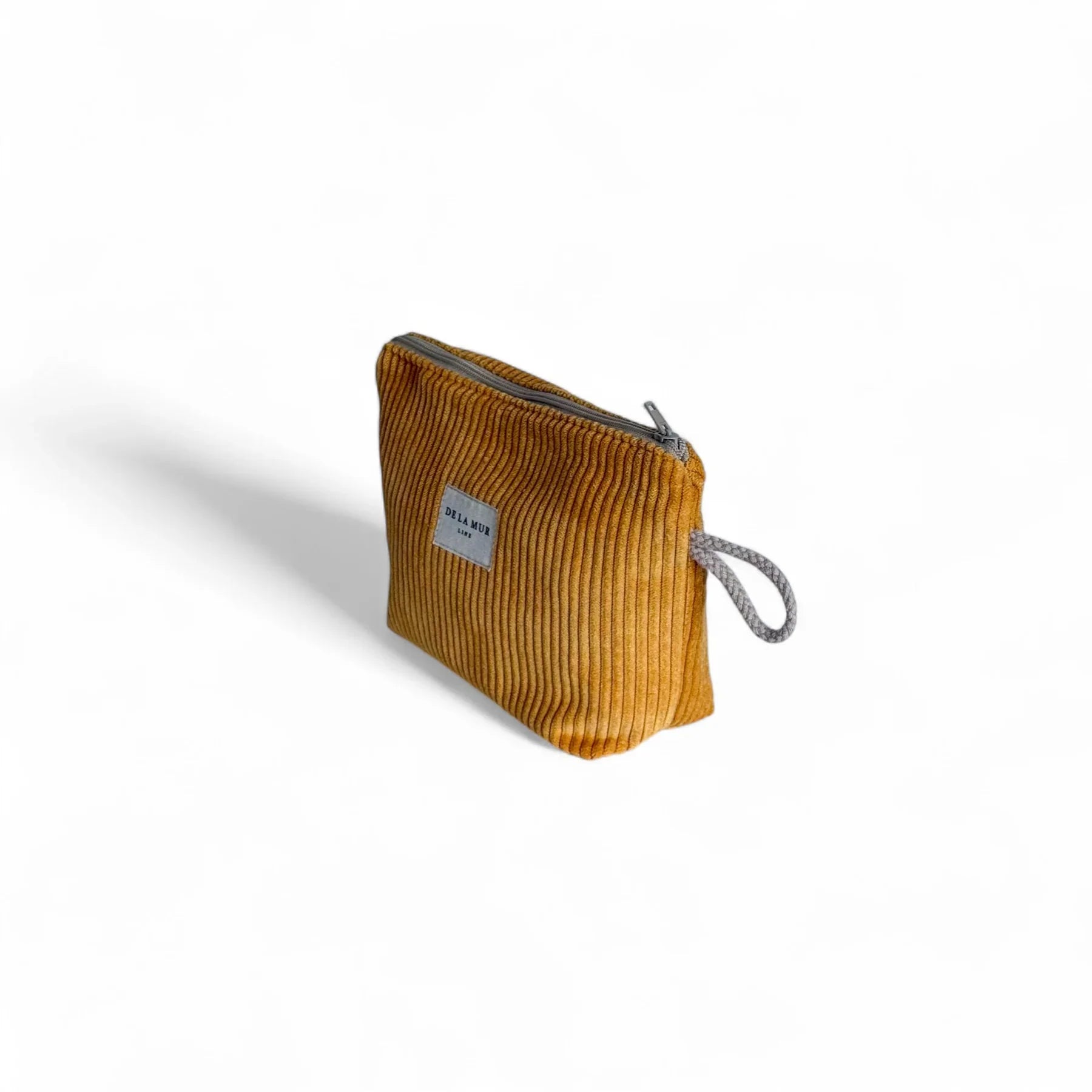 Eine kleine, senfgelbe Cord-Tasche steht im Dreiviertelwinkel auf weißem Hintergrund. Oben befindet sich ein grauer Reißverschluss, seitlich eine graue Schlaufe, vorne das weiße DE LA MUR Label.