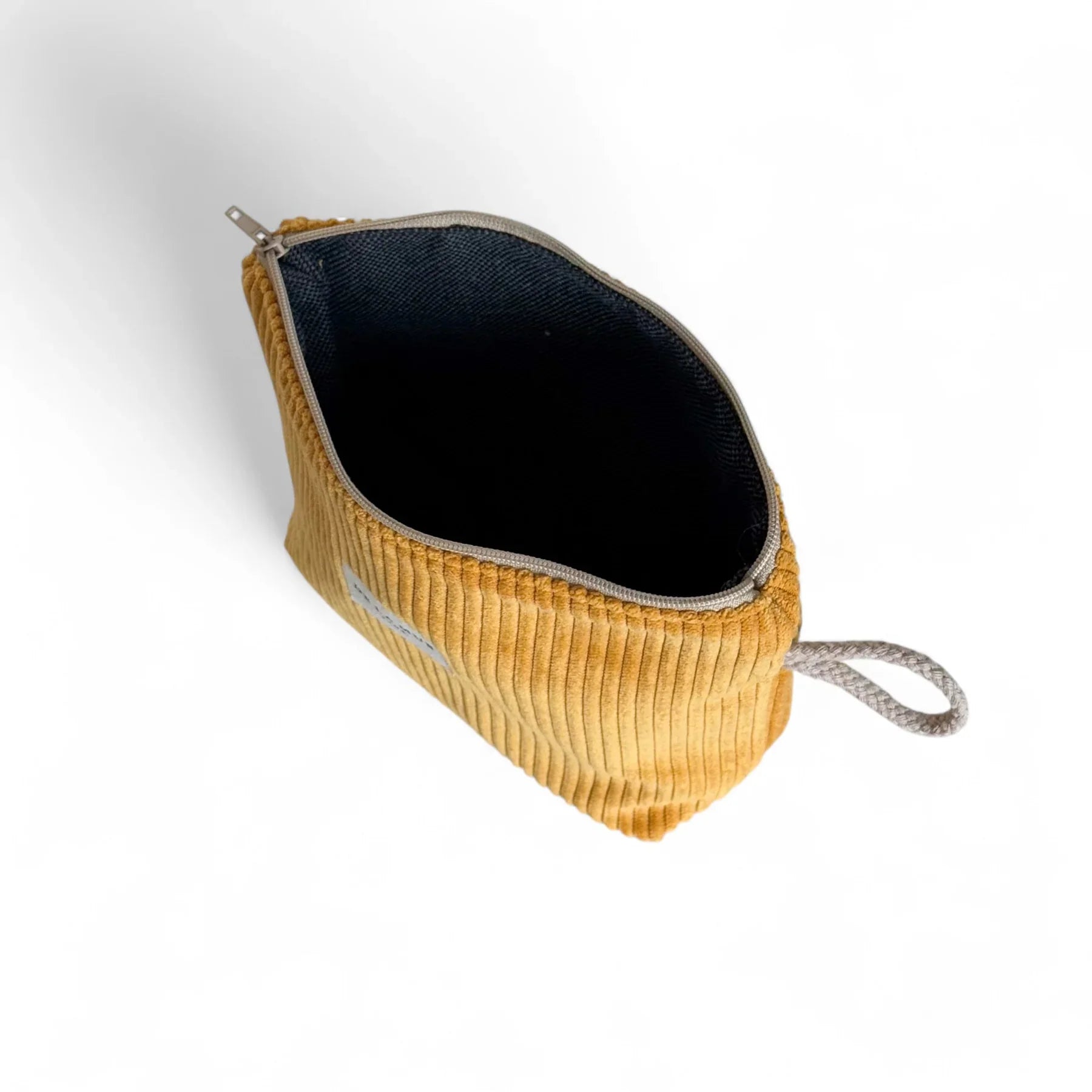 Eine geöffnete, senfgelbe Cord-Tasche mit sichtbarer Rippenstruktur steht auf weißem Hintergrund. Das Innere ist dunkel gefüttert, der Reißverschluss ist geöffnet, seitlich ist eine graue Stoffschlaufe zu erkennen.
