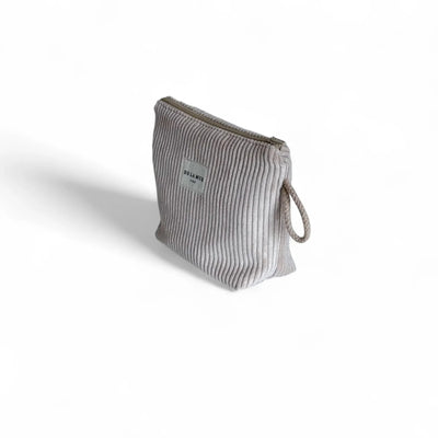 Eine kleine, taupefarbene Cordtasche steht im Dreiviertelwinkel auf weißem Hintergrund. Ein grauer Reißverschluss verläuft oben, seitlich hängt eine kurze geflochtene Schlaufe, vorne ist ein weißes DE LA MUR Label zu sehen. Idealer Organizer.