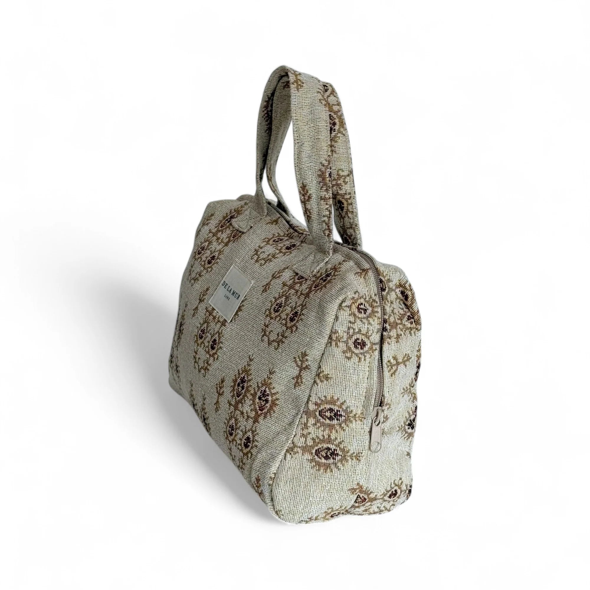 Beigefarbene Beauty Bag aus Jacquard-Stoff mit braunem Ornamentmuster. Die Tasche ist rechteckig, hat zwei Tragegriffe, einen Reißverschluss und zeigt ein helles Label mit der Aufschrift „DE LA MUR Line“. Das Innenfutter ist dunkelgrau.