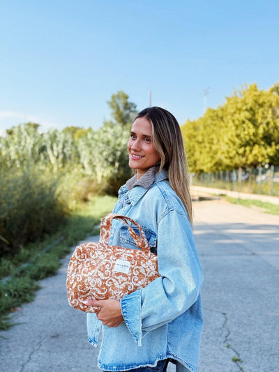 Eine Frau hält eine große Kulturtasche aus rostbraunem Jacquard-Stoff mit Ornamentmuster in der Hand. Die Beauty Bag hat zwei Tragegriffe, ein Innenfutter und ein Label mit der Aufschrift „DE LA MUR LINE“.