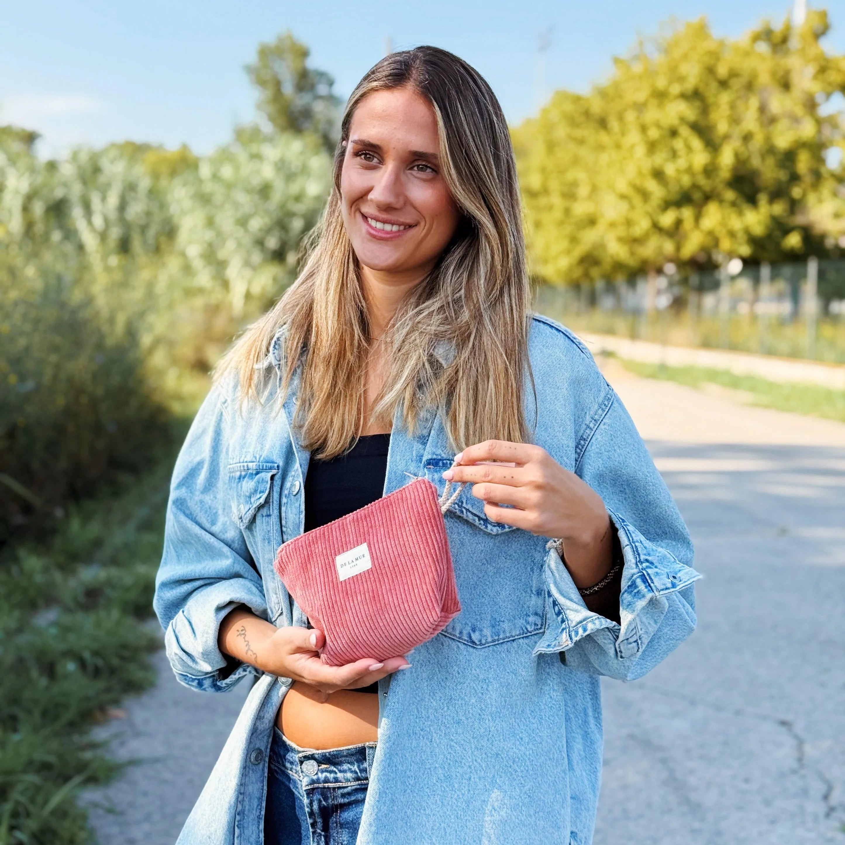 Eine lächelnde Frau mit langen blonden Haaren und Jeansjacke steht draußen auf einem Weg und hält eine kleine korallfarbene Cord-Tasche mit DE LA MUR Label in beiden Händen.