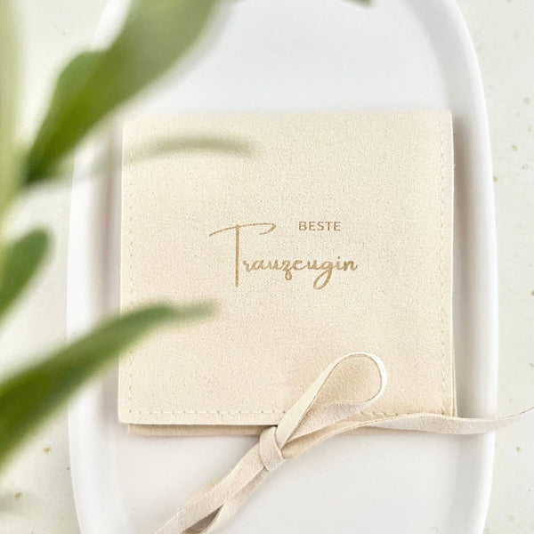 Beigefarbener Schmuckbeutel mit goldener Aufschrift „Beste Trauzeugin“ und Schleifenband, liegend auf einem weißen Teller – Geschenkverpackung zum Trauzeugin-Fragen oder für kleine Schmuckstücke.